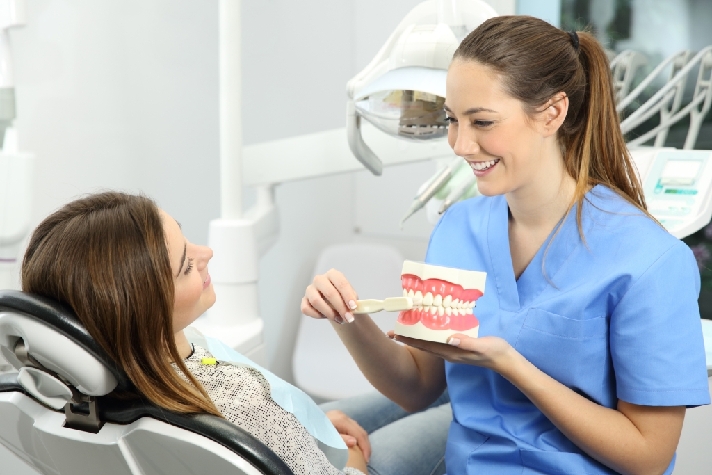 Dentist explaining how to brush teeth correctly to a patient after treatments sitting on a chair in a clinic box with medical equipment in the background